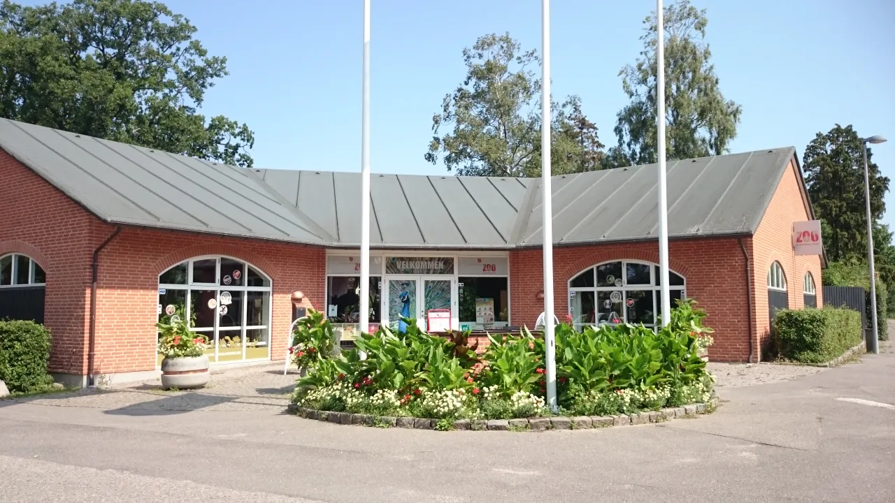 Eingang des Guldborgsund Zoo in Nykøbing Falster: Backstein-Pavillon mit „Velkommen“-Schild und Blumenrabatten.